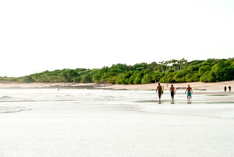 three surfers with surfboards walking down a beach in Costa Rica in the distance