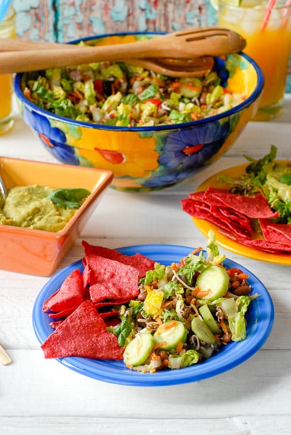 Side salad portion of Taco Salad Ole on blue plate with red tortilla chips and large bowl of salad in the background