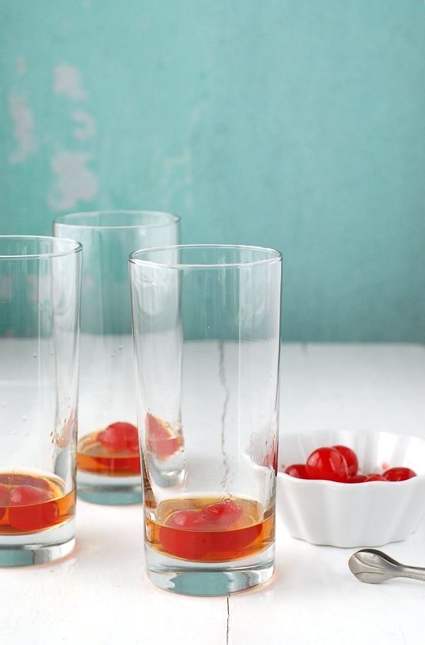 three Collins glasses with bourbon and maraschino cherries with a white bowl of cherries