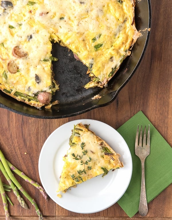 Cooked potato frittata in a cast iron pan with a slice on a white plate placed on a brown wooden table