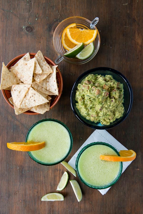 Honeydew Melon Margaritas with orange slices, lime wedges, bowl of chips, freshly made guacamole on a vintage wooden surface