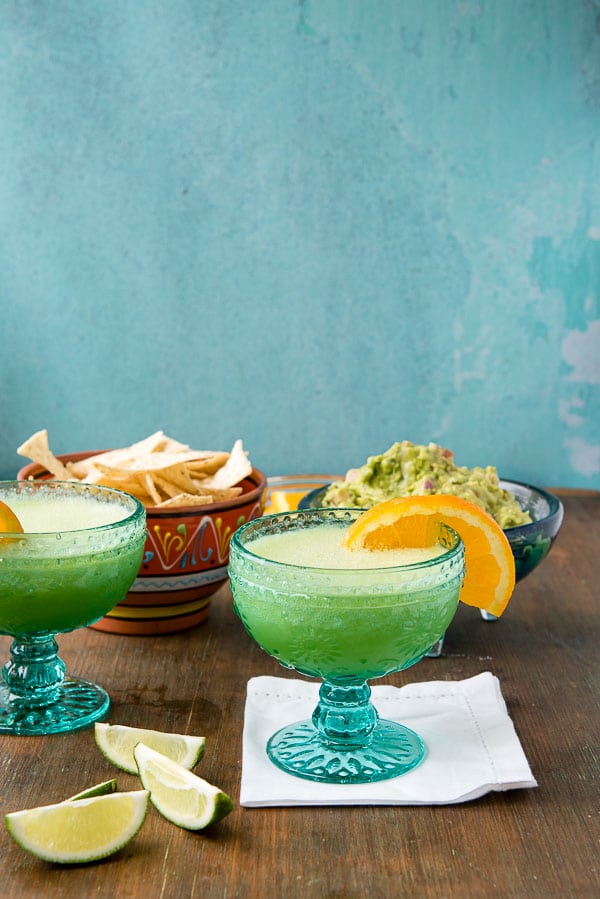 Two Honeydew Melon Margaritas with orange slices on the glass rim, lime wedges, chips in the background