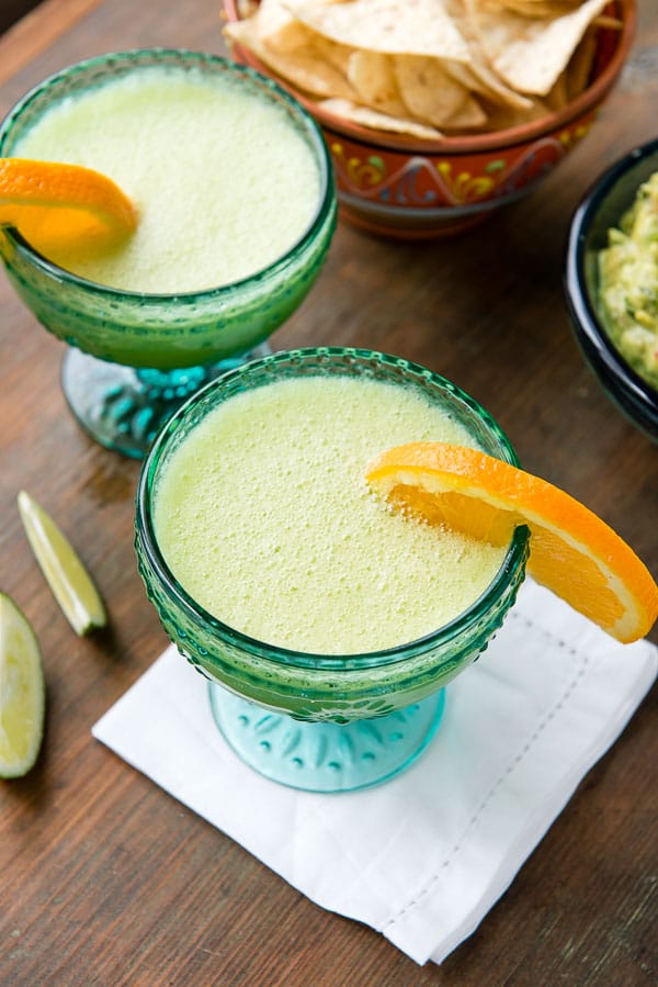 Blue embossed glasses of Honeydew Melon Margaritas with orange slices and guacamole in the background