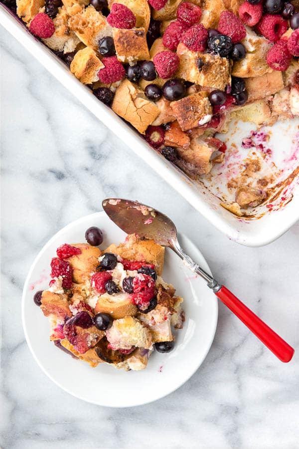 A single serving of Overnight French Toast Casserole with berries on a white plate with a red handled spoon
