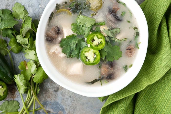 overhead photo of a white bowl filled with Slow Cooker Thai Chicken Mushroom Coconut Soup, garnished with fresh herbs and slices of jalapeno