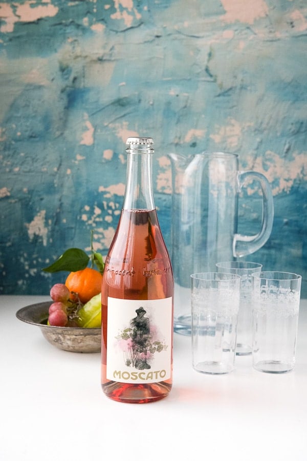 bottle of moscato wine, empty pitcher, and tall glassware, alongside a bowl of fresh fruit to make Moscato Sangria cocktails