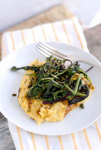 white plate of Pan Browned Polenta with Sauteed Braising Greens and Garlic Scapes sitting on a yellow striped kitchen towel