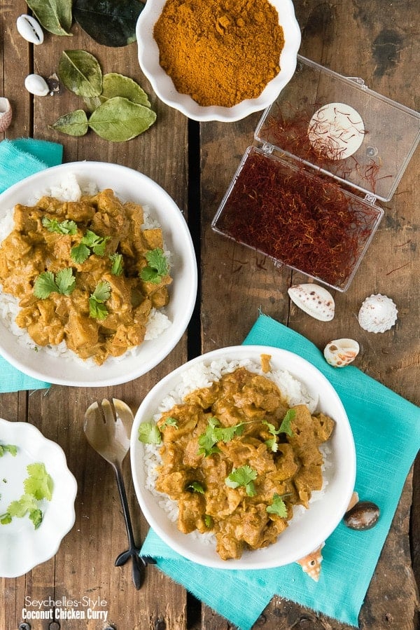 2 white plates of Seychelles-style Coconut Chicken Curry surrounded by some of the ingredients used to make it