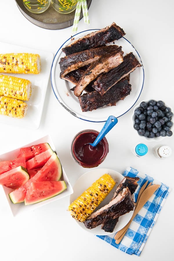 overhead image of a picnic dinner with grilled ribs, corn on the cob, watermelon, and a jar of homemade barbecue sauce