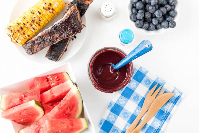 picnic table setting with fresh watermelon slices, corn on the cob, spare ribs, blueberries, and a jar of homemade barbecue sauce