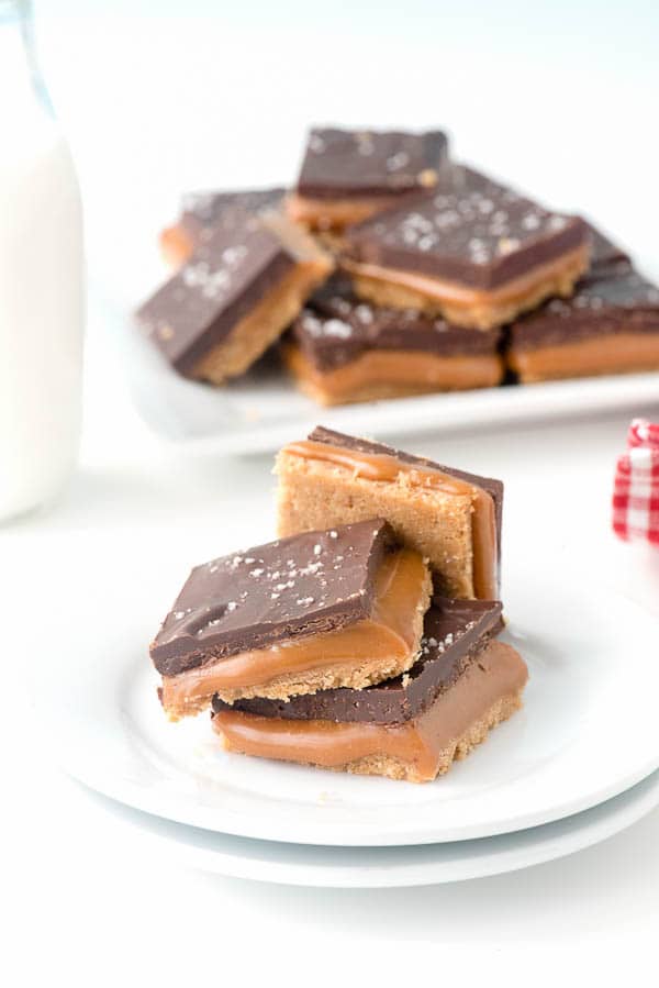 3 No Bake Gluten-Free Millionaire Shortbread recipe bars stacked on a dessert plate. A tray of the no bake chocolate bars is in the background