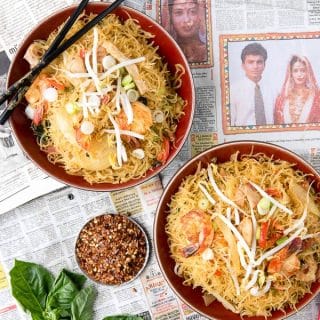 overhead image of 2 bowls of Singapore Street Noodles sitting on top of a Singapore newspaper. Black chopsticks perched on one bowl