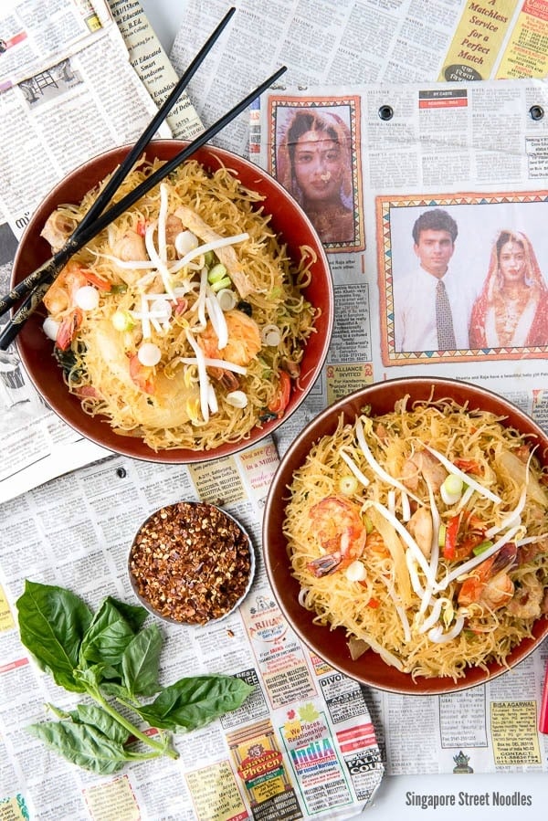 overhead image of 2 bowls of Singapore Street Noodles sitting on top of a Singapore newspaper. Black chopsticks perched on one bowl