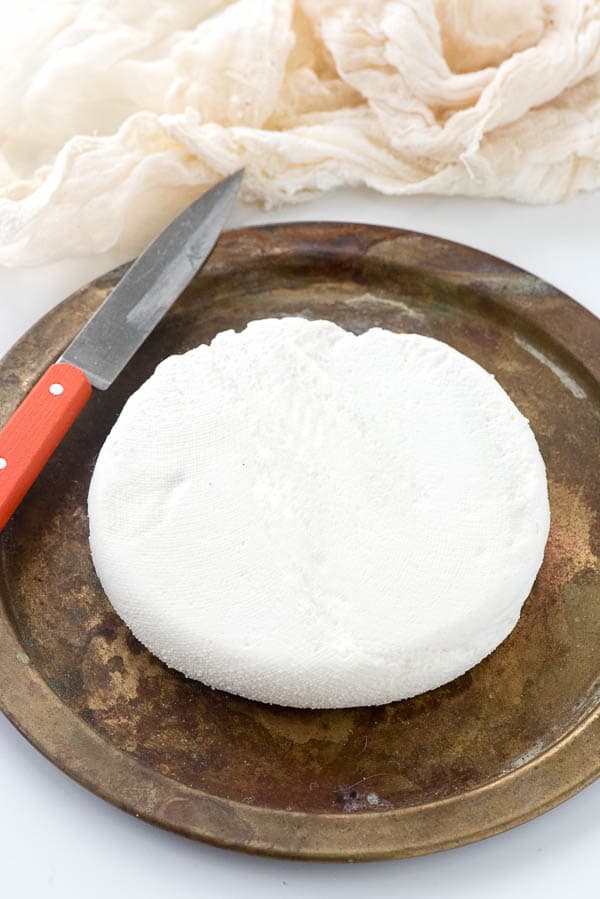 a wheel of Homemade Paneer cheese on a wooden cheese board with a knife for slicing sits next to a piece of cheesecloth