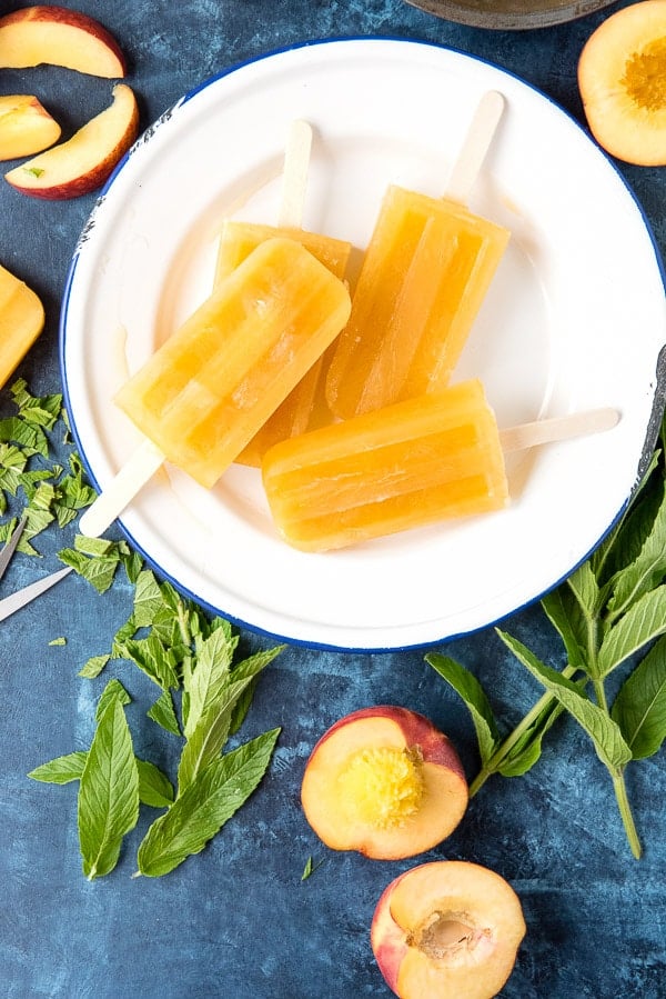 frozen peach popsicles flavored with ginger and mint sitting on a white plate, surrounded by the ingredients used to make them