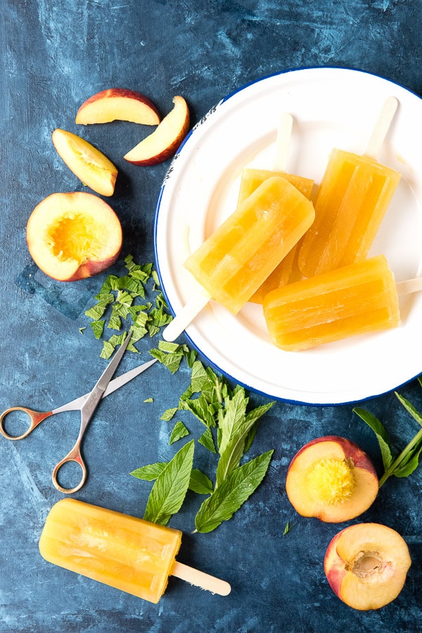 a white plate stacked with frozen peach popsicles, surrounded by fresh peaches and snipped mint leaves