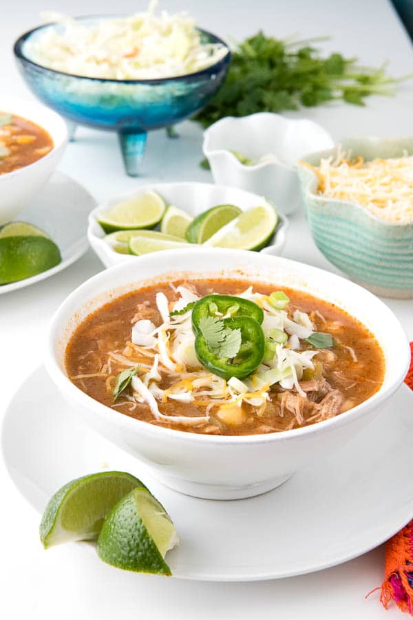 white soup bowl filled with red posole soup made in a slow cooker sitting on a white plate with lime wedges