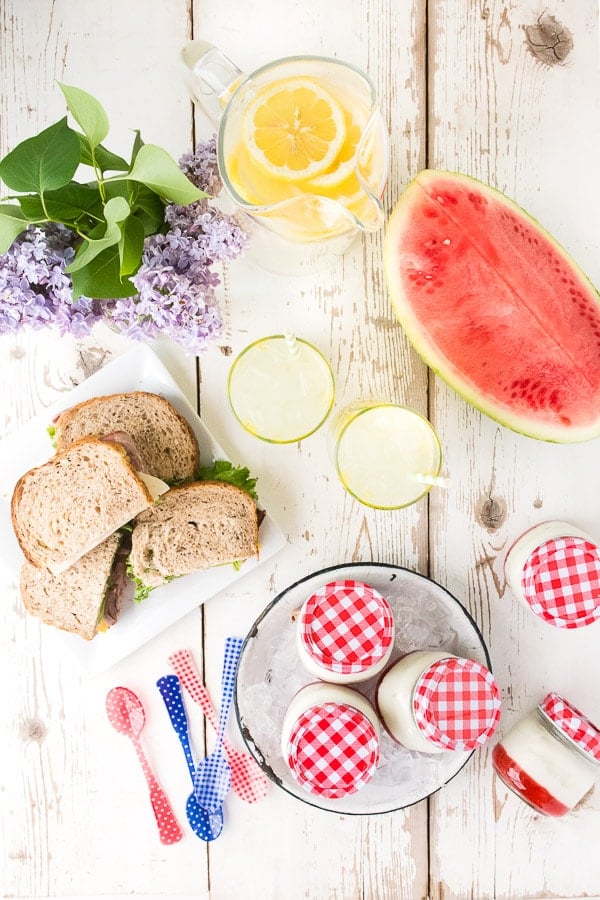 overhead image of a white wooden picnic table arranged with sandwiches, watermelon, lemonade, and portable panna cotta desserts made with strawberry jam