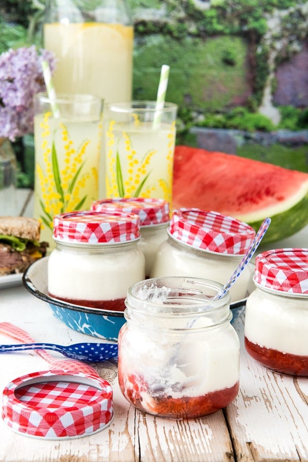 jars of homemade Panna Cotta with strawberry jam sitting on a picnic table with watermelon and glasses of lemonade