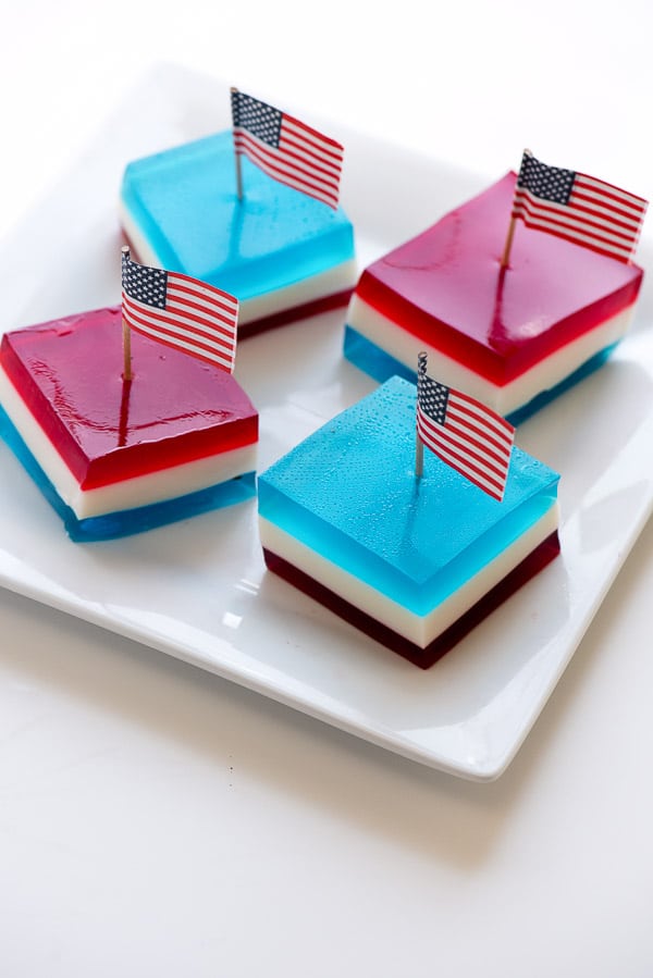 Four squares of red white and blue layered finger jello on a white plate with American flag picks on top