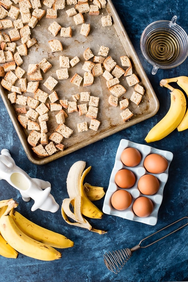 ingredients for bananas foster bread pudding (cubed bread on a rimmed baking sheet, peeled bananas, cream)