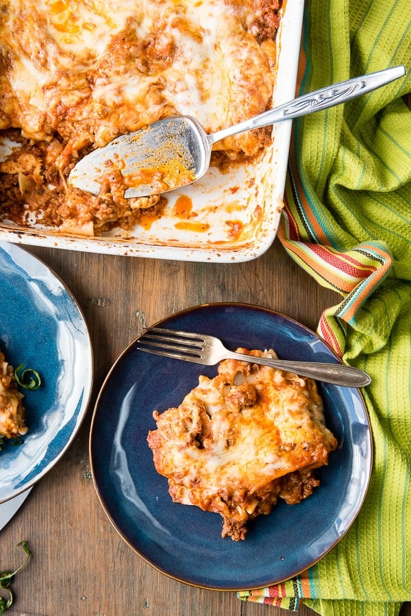 serving of meat lasagna on blue plate with part of white lasagna dish showing with serving scooped out