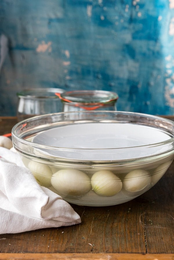 side view of pearl onions being held down in glass bowl of brine by a small plate