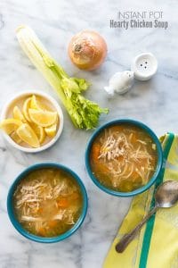 Two blue bowls of hearty chicken soup with a bowl of lemon wedges, fresh celery stalks, and onion and green napkin