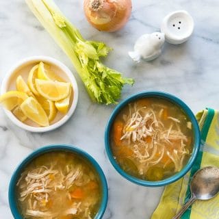 Two blue bowls of hearty chicken soup with a bowl of lemon wedges, fresh celery stalks, and onion and green napkin