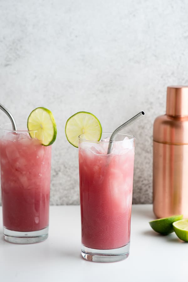 Two red Cape Cod Cooler cocktails in tall glasses with ice, lime slices, metal straws 