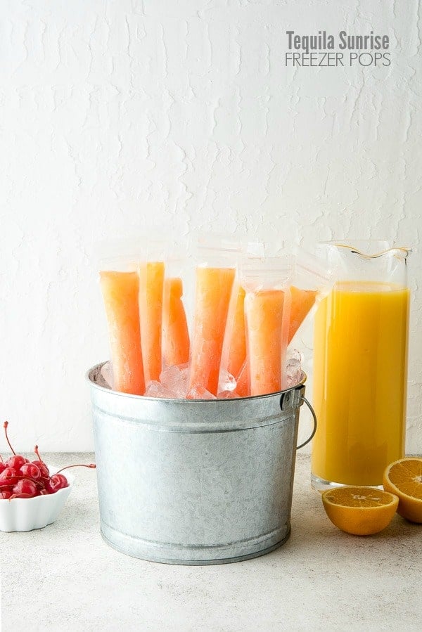Boozy orange Tequila Sunrise Freezer Pops in a mini galvanized bucket with a glass pitcher of orange juice