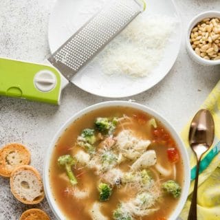 White bowl of Pesto Tortellini Soup with toasted baguette slices, grated Parmesan cheese