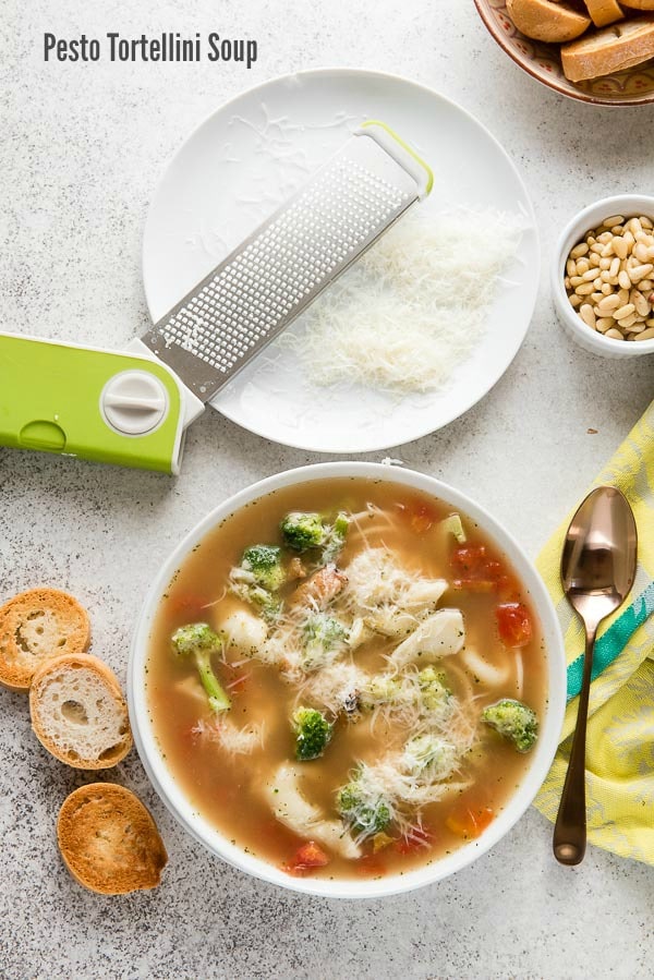 White bowl of Pesto Tortellini Soup with toasted baguette slices, grated Parmesan cheese