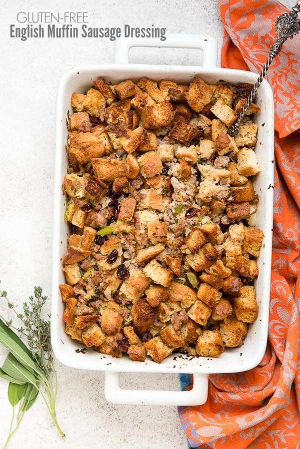 White porcelain baking dish of Gluten-Free English Muffin Sausage Dressing with orange kitchen towel and fresh herb