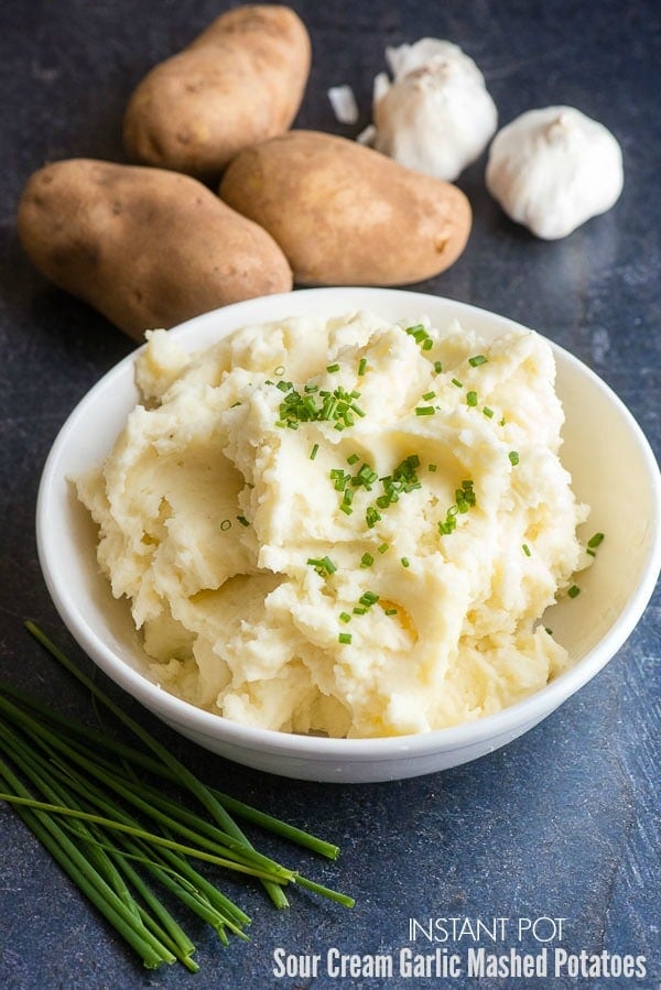Instant Pot Sour Cream Garlic Mashed Potatoes in a white bowl with fresh chives, russet potatoes and garlic on a slate stone surface