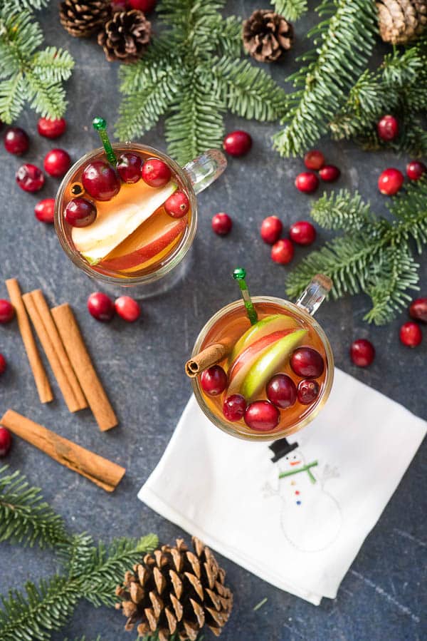  Two steaming hot glass mugs of Cranberry Apple Hot Toddy cocktail with evergreen boughs, cranberries and cinnamon sticks