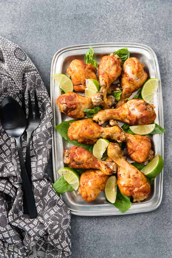 A pewter platter of Instant Pot Smoked Paprika & Garlic Chicken Legs with lime wedges, black serving utensils and gray kitchen towel