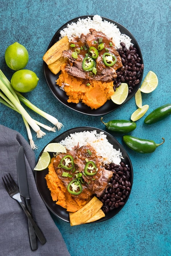 Two plates of Slow Cooker Pot Roast with spicy sauce, jalapeno slices, mashed sweet potatoes, rice, beans and fried plantains