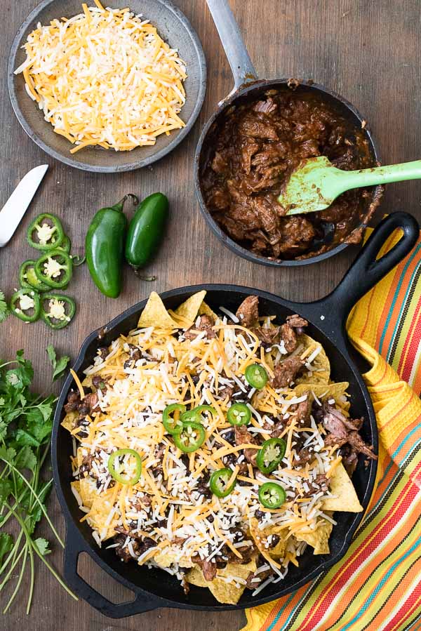 Pot Roast Nachos ready for the oven with two types of grated cheese, pot roast in sauce, sliced jalapenos in a black cast iron skillet