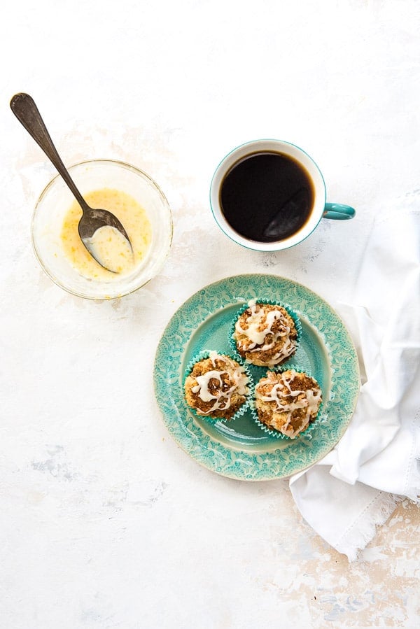 Three Coffee Cake Muffins on a blue plate with a cup of coffee and small bowl of fresh orange glaze with vintage spoon