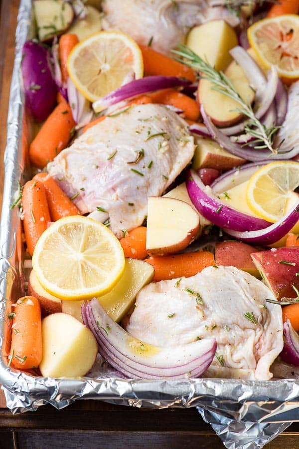 Fresh chicken thighs, baby carrots, potatoes, red onion wedges, lemon slices and rosemary sprigs on a sheet pan