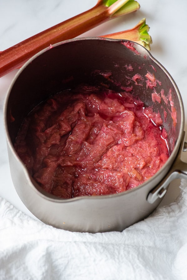 Sweetened cooked rhubarb in a taupe saucepan with rhubarb stalks in the background