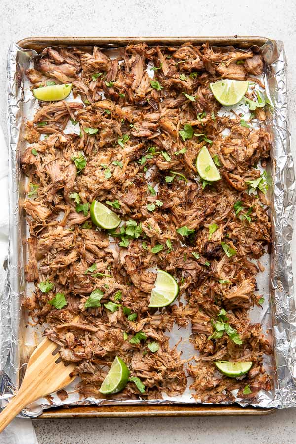 Sheet pan of cooked Pineapple Citrus Slow Cooker Carnitas with cilantro leaves and lime wedges