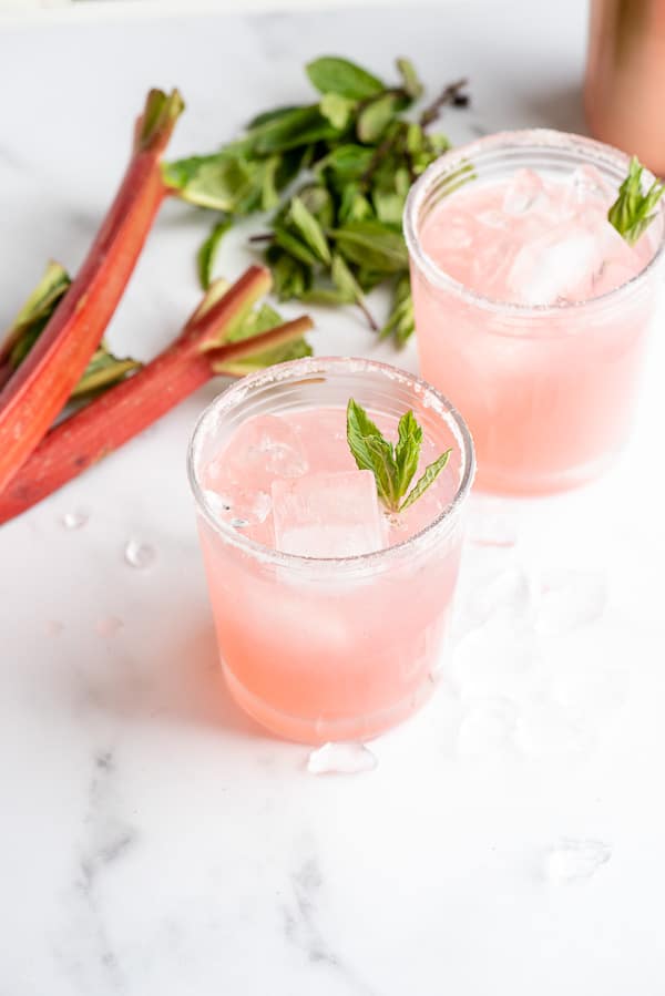 Two light pink Rhubarb Pie Cocktails (a rhubarb vodka cocktail) with rhubarb stalks in background