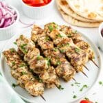white plate of grilled Chicken Souvlaki skewers with flatbread and vegetables in the background