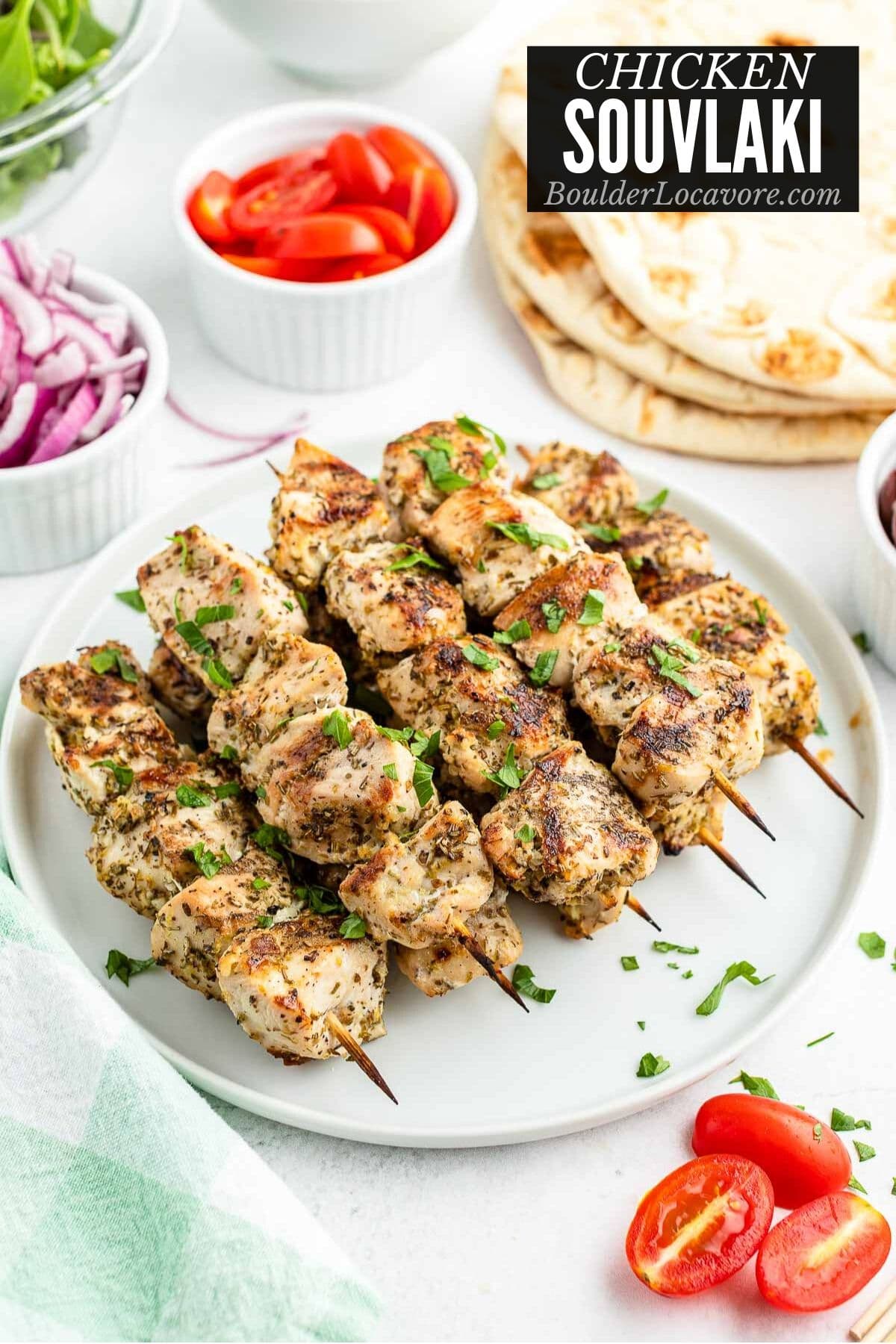 white plate of grilled Chicken Souvlaki skewers with flatbread and vegetables in the background