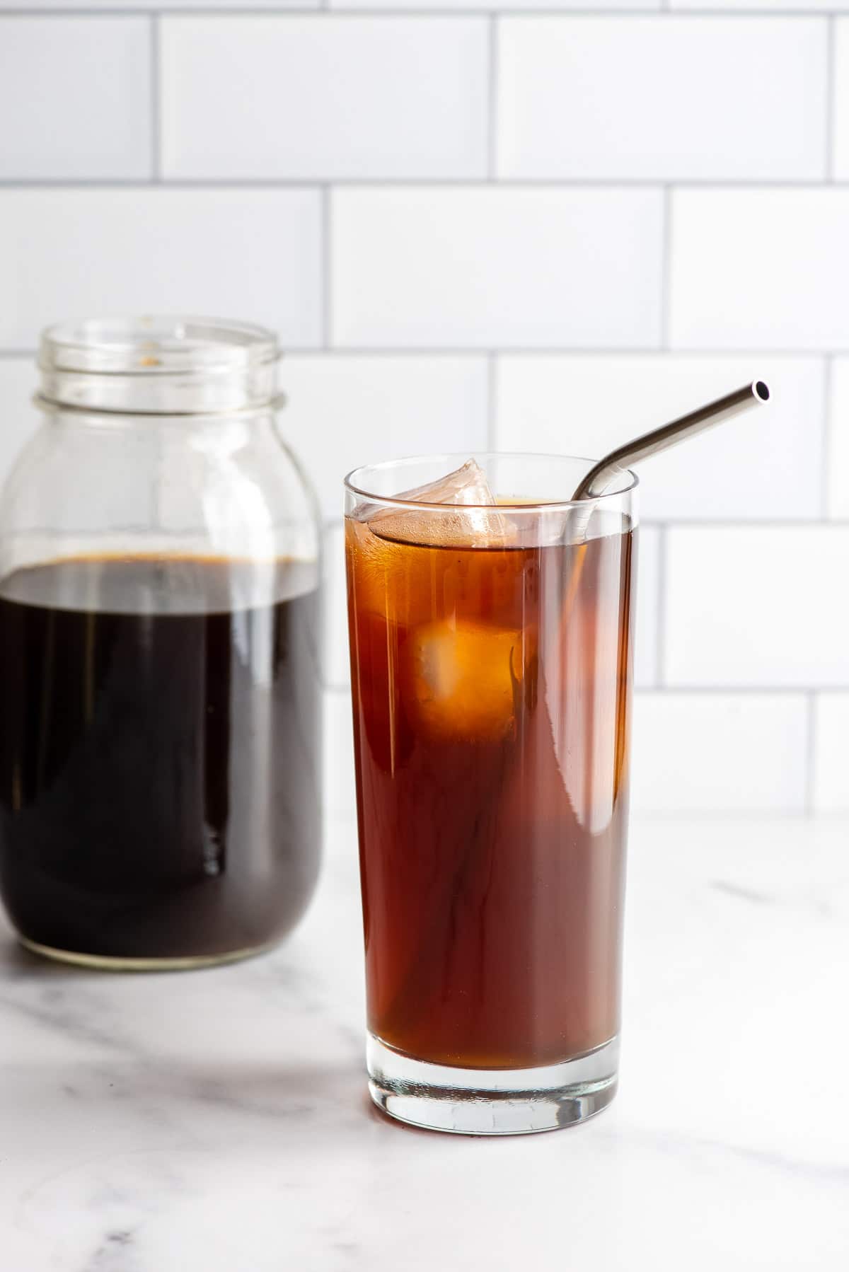 glass of cold brew coffee with ice and a metal straw and jar of coffee behind