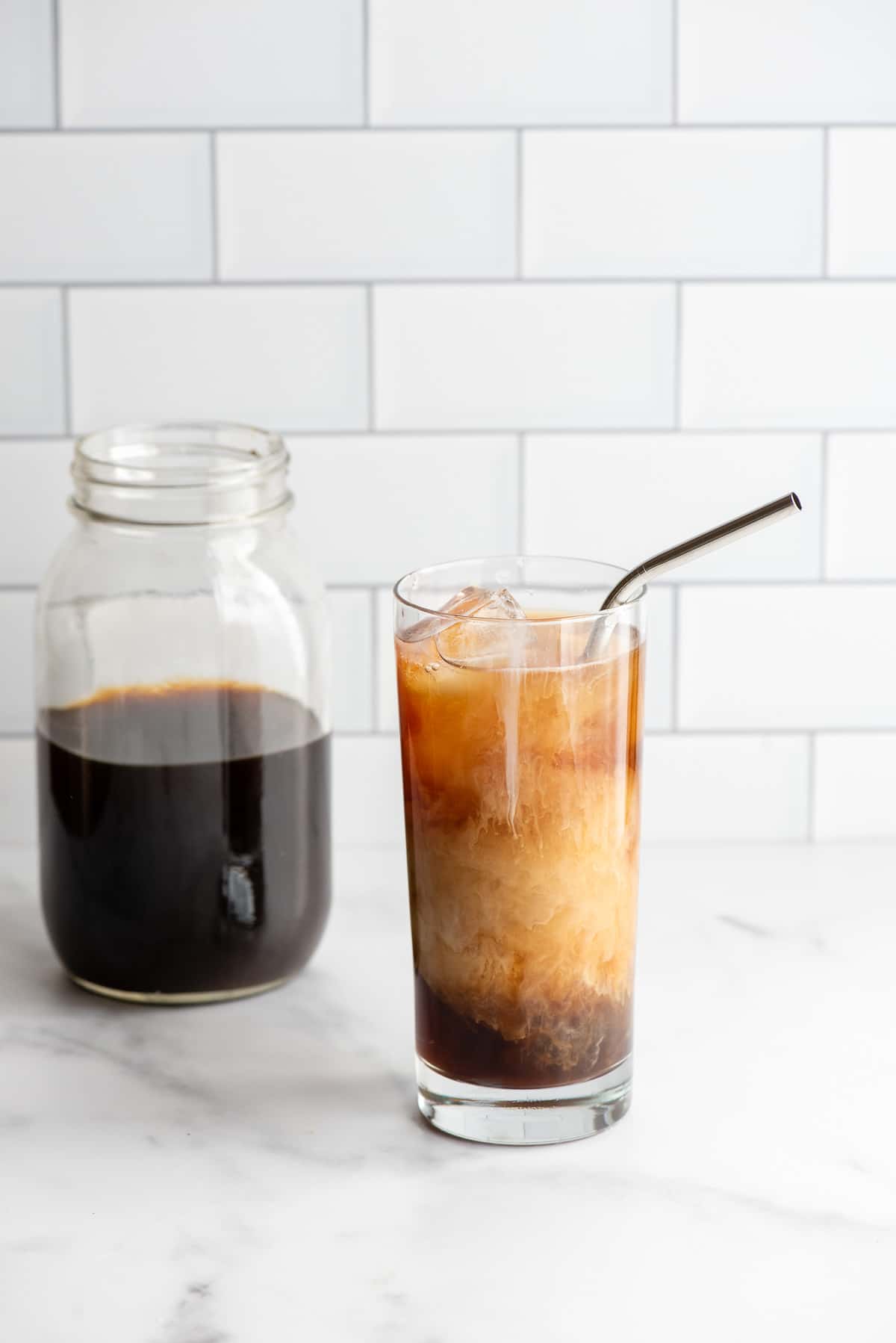 glass of cold brew coffee with cream and a metal straw with a jar of cold brew coffee in a jar in background